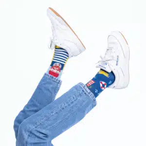 Person wearing white trainers, blue jeans, and mismatched Fisher's Tale socks—one featuring a cartoon pirate, the other nautical symbols—shows off curated Estonian design while posing with legs crossed against a plain white background. Gate21.ee