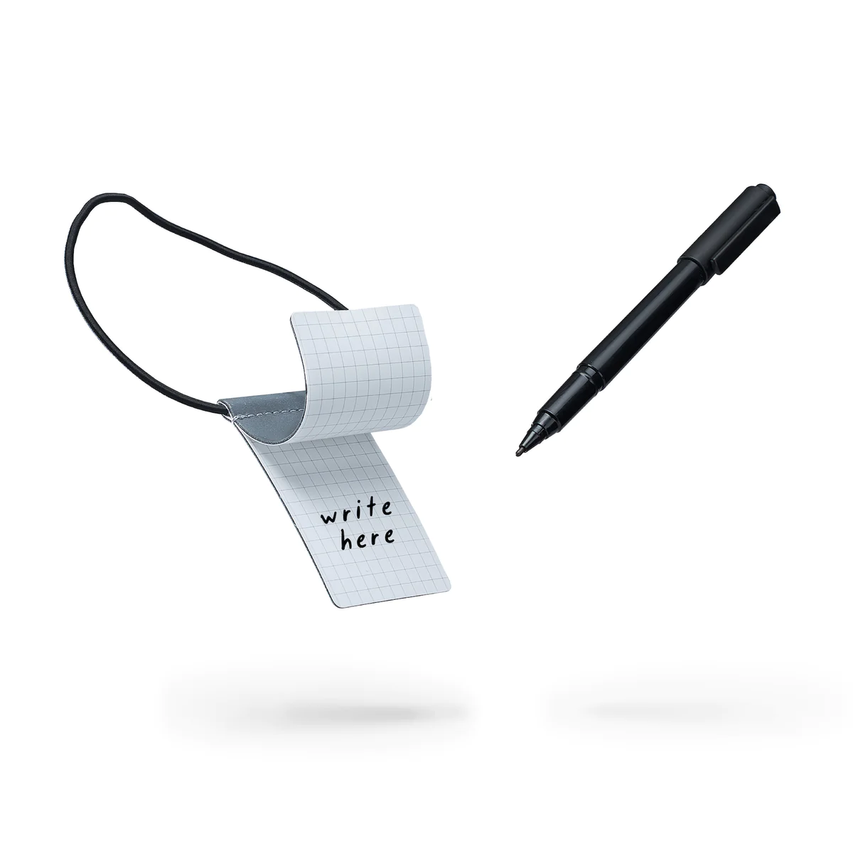 The Luggage Tag - Black features a small gridded notepad reading "write here," attached by a black elastic band. A matching black pen completes the set, both displayed against a white background&mdash;ideal for Tallinn souvenirs or curated gift shops. Gate21.ee