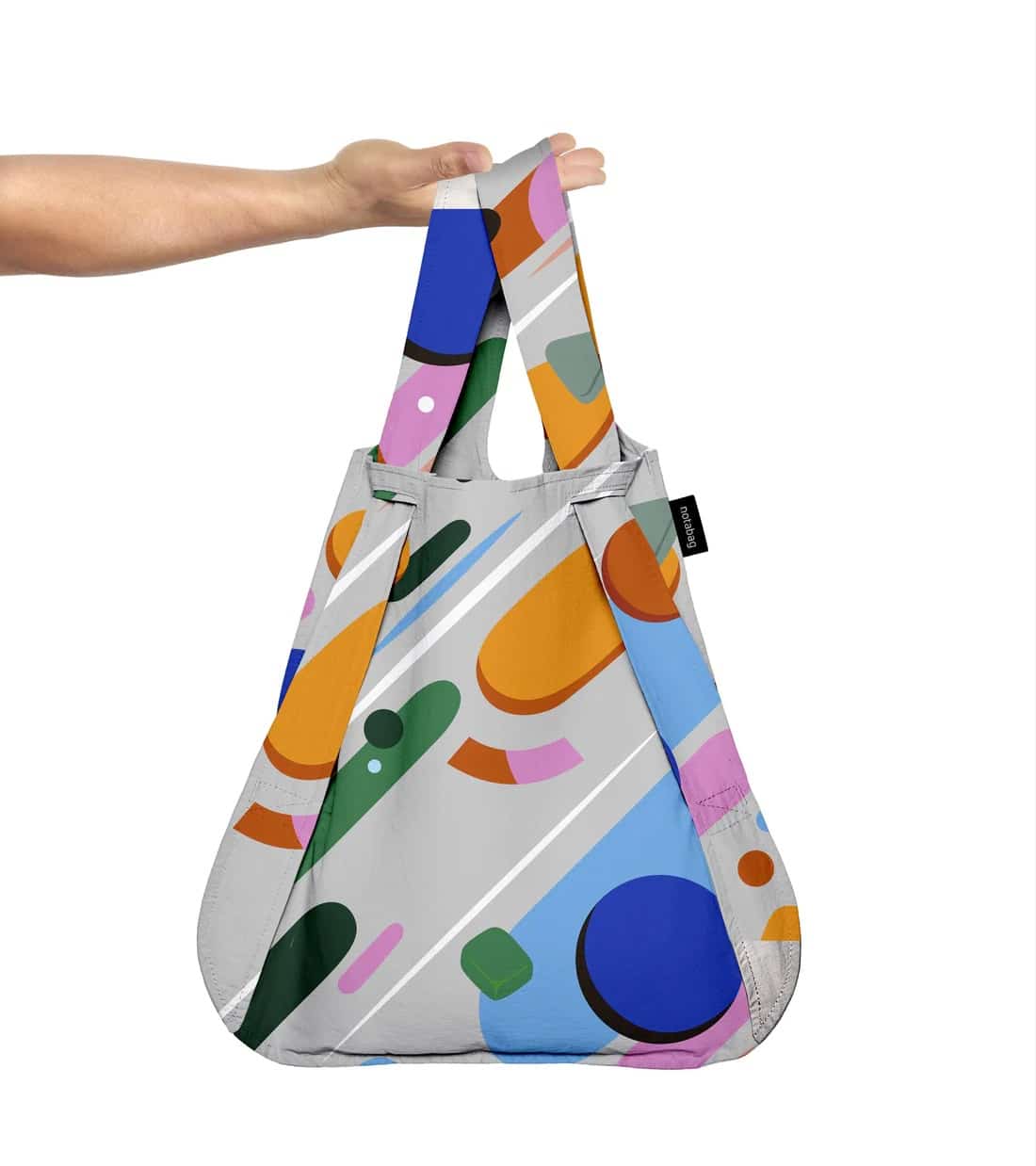 A hand holds the Fruit Salad - Notabag, a reusable shopping bag with colourful abstract shapes and Estonian design flair. It features a short handle, black side tag, and light grey background—ideal for any concept store. Gate21.ee