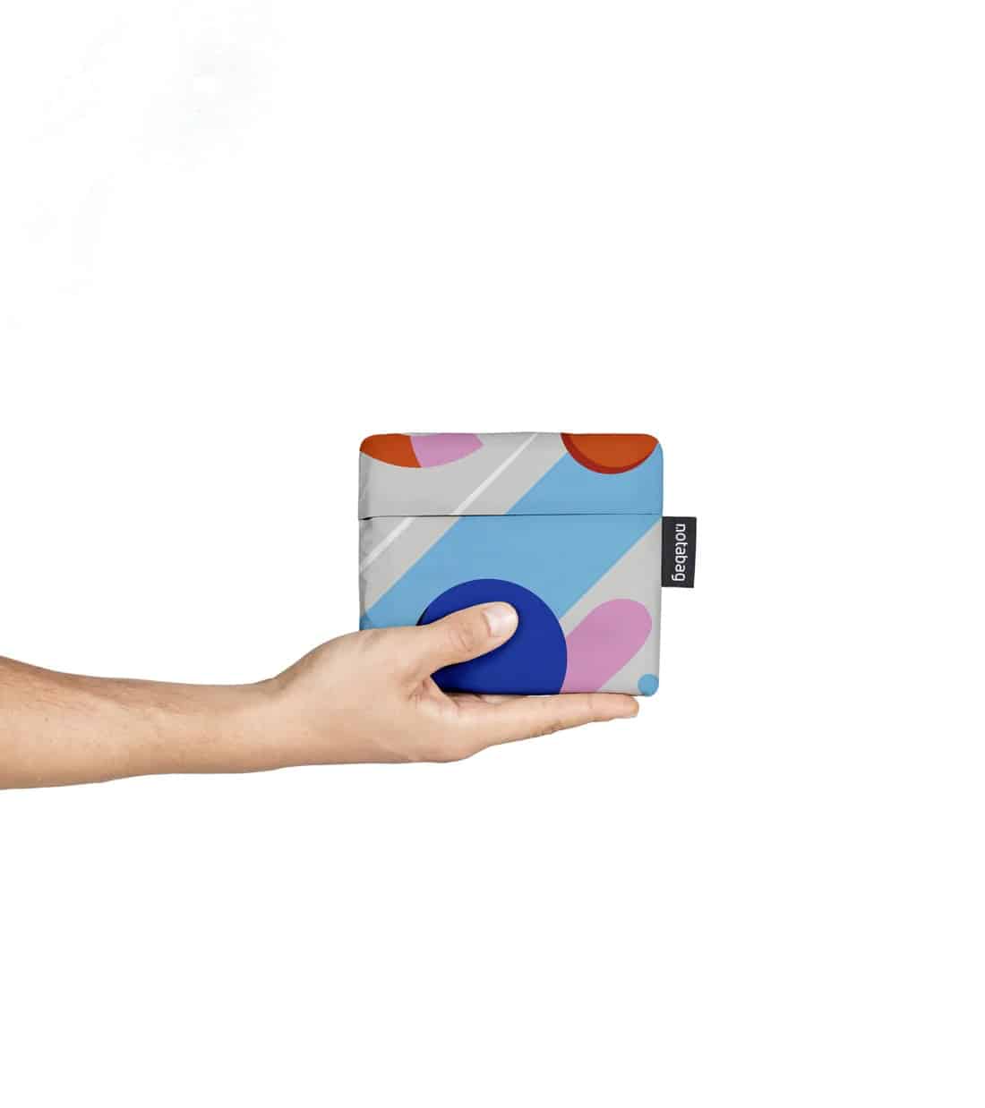 A hand holds the Fruit Salad - Notabag, a small, rectangular item with a vibrant abstract design in blue, purple, orange, and grey—highlighting Estonian aesthetics. A black tag with white text accents the side. The background is white. Gate21.ee