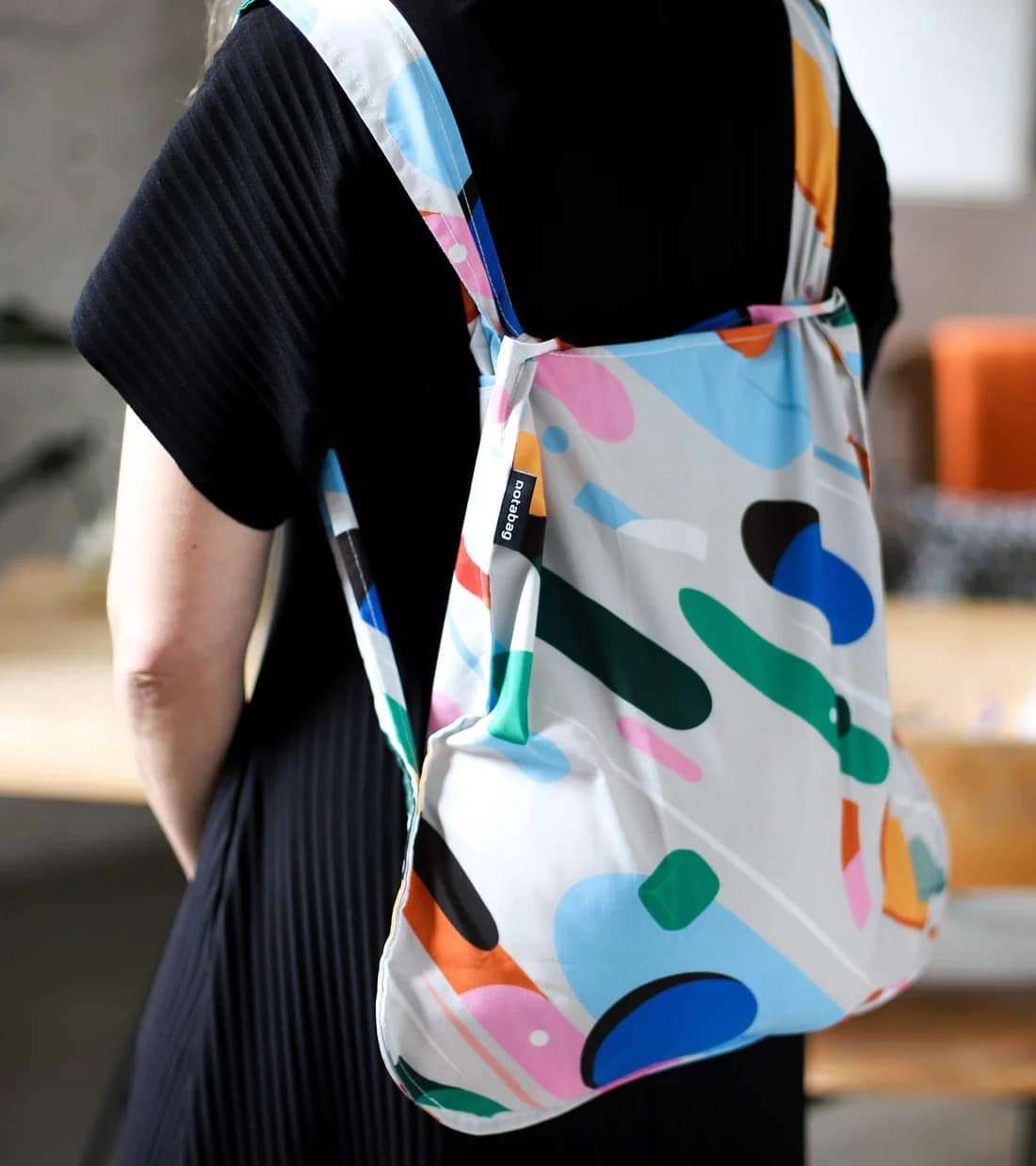 A person in a black pleated dress carries the Fruit Salad - Notabag, a colourful abstract-patterned fabric rucksack with Estonian design and brand tag. A blurred table and orange chair in the background evoke Tallinn’s shopping charm. Gate21.ee