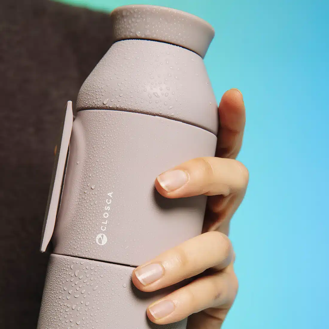 A hand holds the Closca Wave - Himalaya water bottle, light grey with a matte, minimalist finish and condensation beads, set against a gradient blue background—a stylish pick for your next Tallinn concept or gift shop visit. Gate21.ee