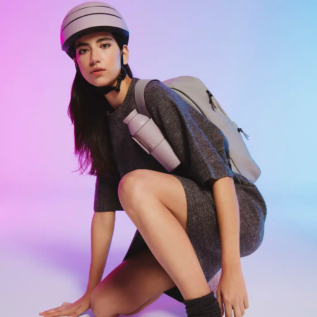 A person with a Closca Wave - Himalaya bottle kneels on the floor against a pink-blue gradient, wearing a grey dress, helmet, and rucksack—embracing the minimalist style seen in modern Estonian design shops. Gate21.ee