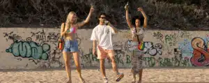 Three people stroll a sandy beach by a graffiti wall, dressed in casual summer wear. One sports the stylish New York Rolltop Rucksack - Olive, perhaps after exploring curated Tallinn shopping or browsing a local concept shop. Gate21.ee