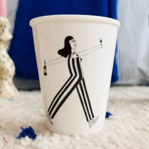 The Wine O'clock Cup is a white cup featuring a black and white illustration of a woman in striped trousers holding a wine bottle and glass. This stylish Estonian design makes it an ideal addition to any gift shop or concept store. Gate21.ee