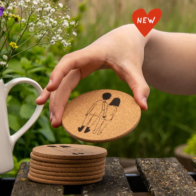 A hand lifts a round Helen B Coaster - KÕIK featuring minimalist art of two people. More coasters are stacked on wood, with a white jug and flowers in the background. A red "NEW" label is at the top right, highlighting Estonian design. Gate21.ee