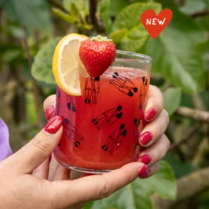 A hand with pink nail varnish holds a Helen B Glass - KÕIK filled with red drink, garnished with lemon and strawberry. There’s a “NEW” red heart at the top right, and green leaves fill the background. Gate21.ee
