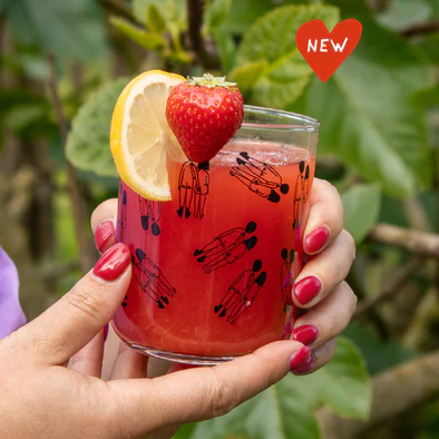 A hand with pink nail varnish holds a Helen B Glass - KÕIK filled with red drink, garnished with lemon and strawberry. There’s a “NEW” red heart at the top right, and green leaves fill the background. Gate21.ee