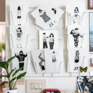 Helen B Postcards – KÕIK: A curated set of black-and-white illustrated postcards with people in various poses, displayed on a white wire rack with a green plant. Perfect for gift shops or fans of unique Estonian design. Gate21.ee
