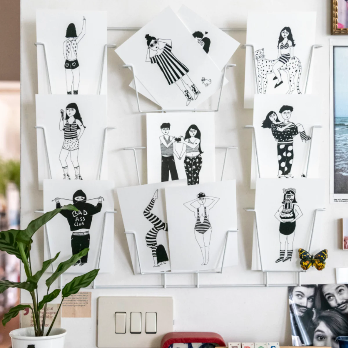 Helen B Postcards – KÕIK: A curated set of black-and-white illustrated postcards with people in various poses, displayed on a white wire rack with a green plant. Perfect for gift shops or fans of unique Estonian design. Gate21.ee