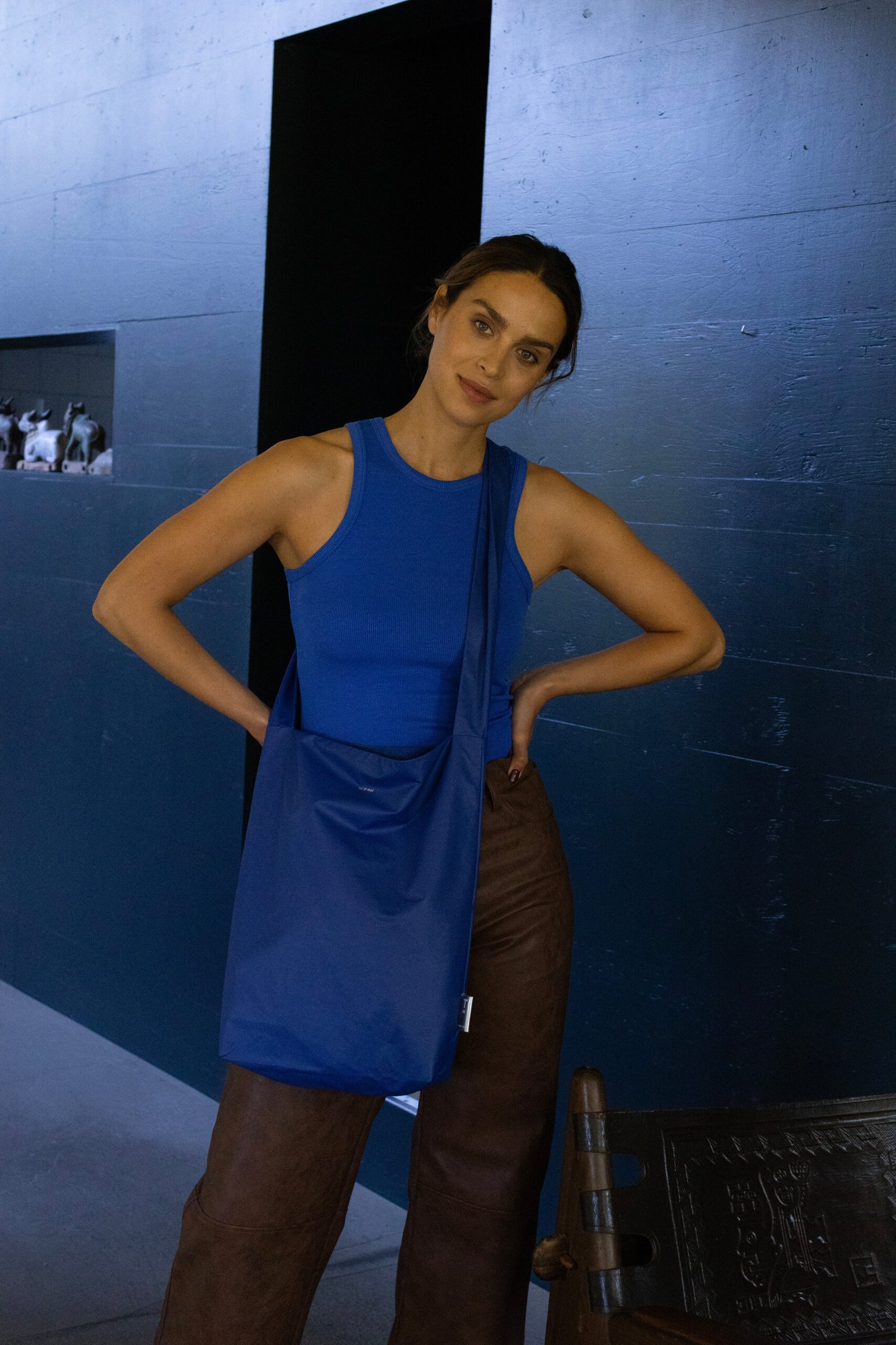 A woman in a blue sleeveless top, brown trousers, and a Feel Good Bag - Sodalite Blue stands indoors with her hands on her hips in front of a dark wall and black doorway. Gate21.ee