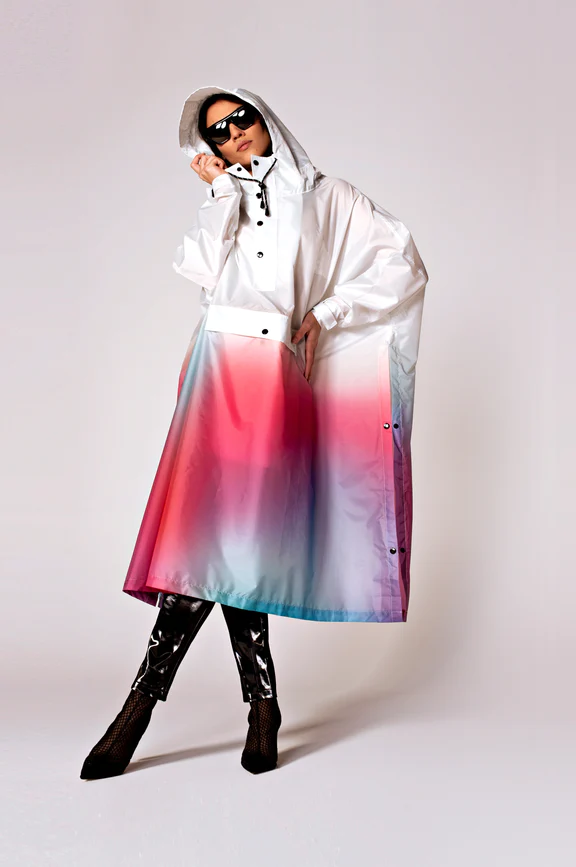 A person wears the glossy, hooded Disco Dream rain poncho by Estonian design with a white top and pink-red-blue gradient skirt. Black sunglasses, leggings, and heeled shoes complete this chic Tallinn shopping look. Gate21.ee