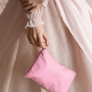 A person wearing a light pink, sheer long-sleeved dress holds a small, zippered Feel Good Bag – Pink Dolphin with one hand resting over the other at waist level. Gate21.ee