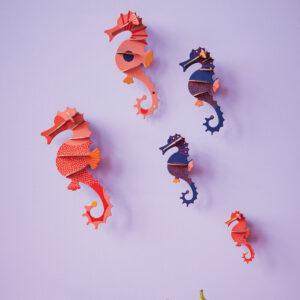 Sea Horses: Five stylised sculptures in red, orange, and blue, featuring curated Estonian design, are mounted on a pale purple wall above an orange surface with peaches and figs below. Gate21.ee