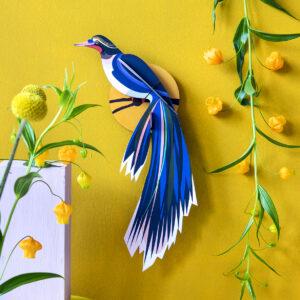 The Paradise Bird Flores, a vibrant blue paper bird with a long tail in Estonian design, is displayed on a yellow wall among yellow flowers and green vines, with a white block and yellow round base in the lower left corner. Gate21.ee