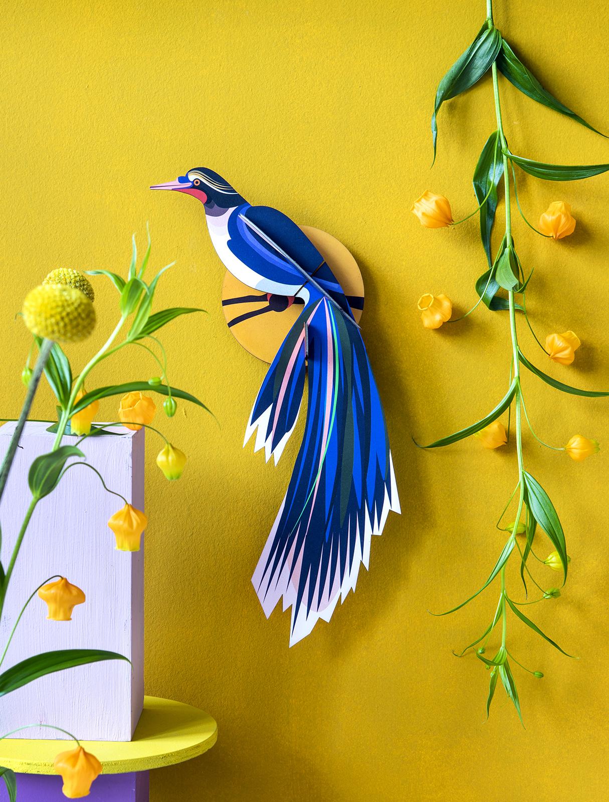 The Paradise Bird Flores, a vibrant blue paper bird with a long tail in Estonian design, is displayed on a yellow wall among yellow flowers and green vines, with a white block and yellow round base in the lower left corner. Gate21.ee