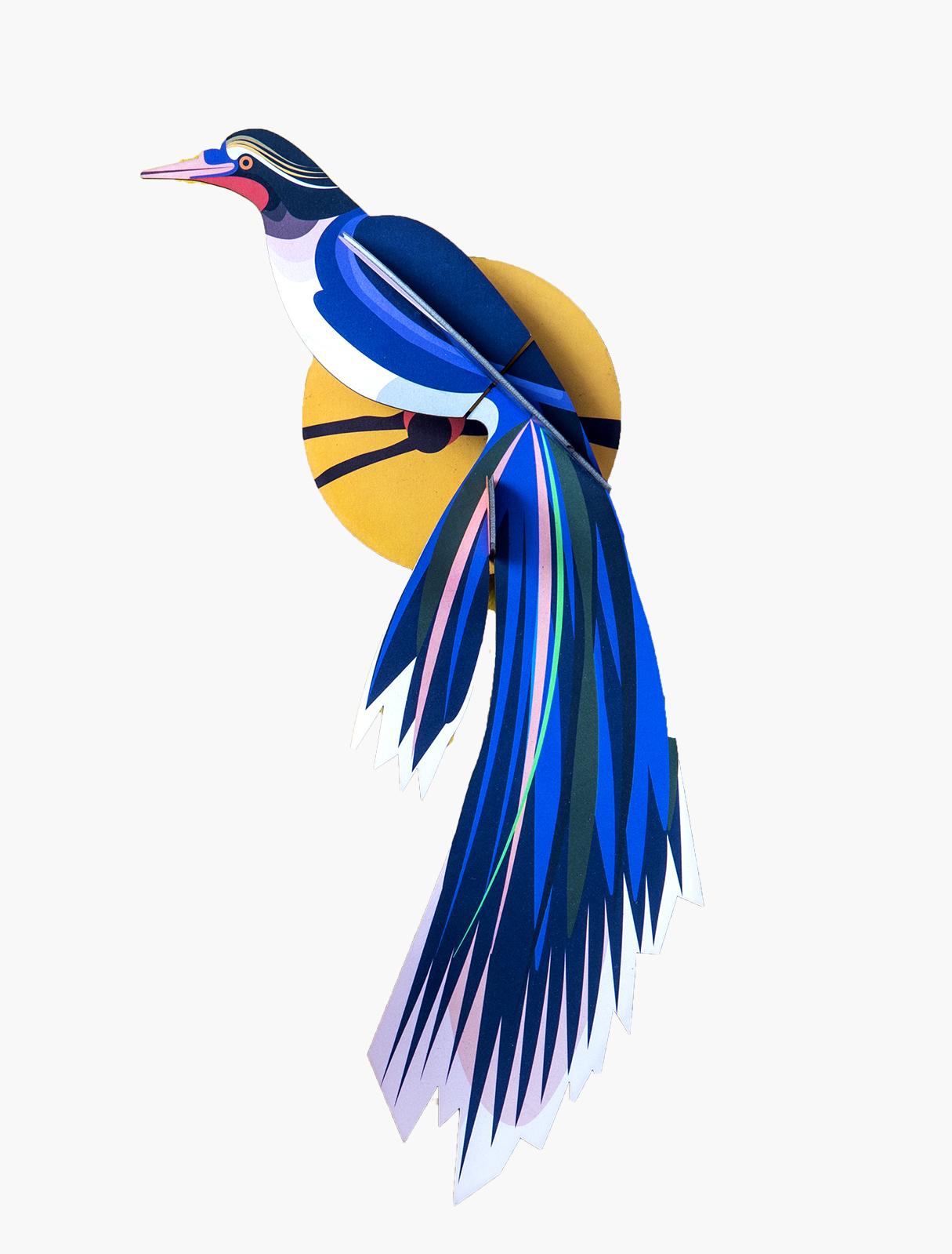 Paradise Bird Flores features a stylised paper bird with blue, purple, and white feathers perched before a yellow circle—an eye-catching piece inspired by Estonian design, perfect for adding unique flair to your gift shop. Gate21.ee