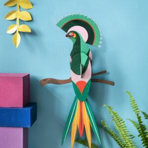 Paradise Bird Gili is a vibrant paper art piece featuring green, orange, pink, and yellow feathers on a brown branch against a bright blue wall—an ideal Tallinn shopping find for unique Estonian design or a thoughtful present. Gate21.ee