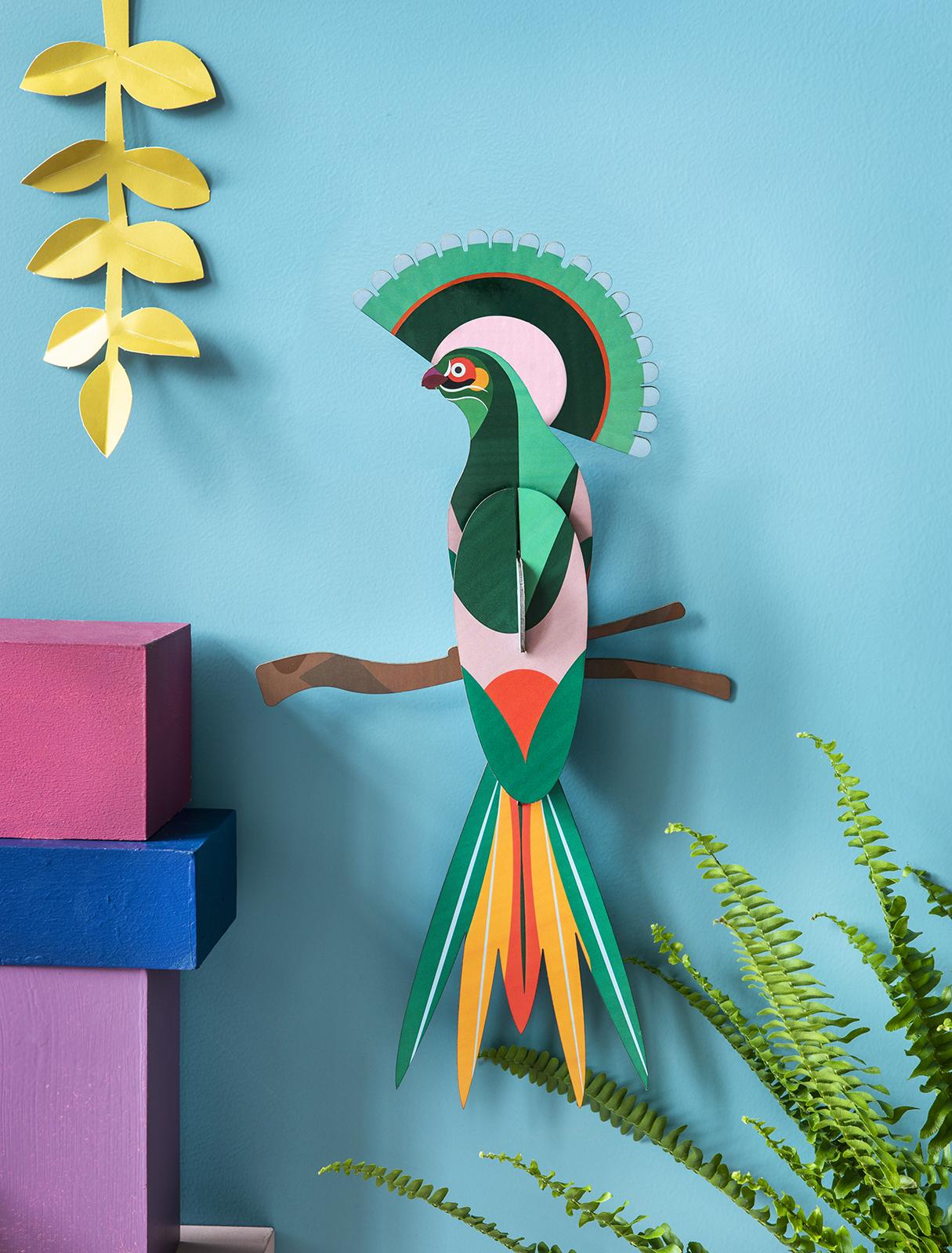 Paradise Bird Gili is a vibrant paper art piece featuring green, orange, pink, and yellow feathers on a brown branch against a bright blue wall—an ideal Tallinn shopping find for unique Estonian design or a thoughtful present. Gate21.ee