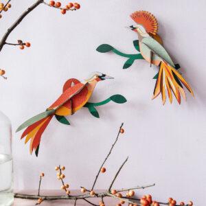 Two Paradise Bird Obi decorations with vibrant orange, yellow, and red feathers brighten a pale pink wall beside berry branches; a glass water jug rests below—fresh inspiration from an Estonian design gift shop. Gate21.ee