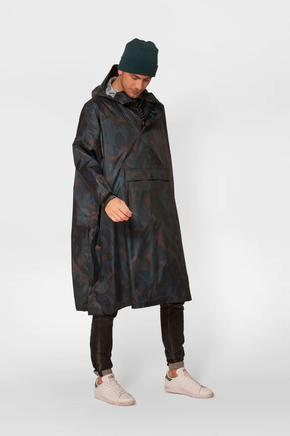 Wearing the Storm Camo rain poncho, black trousers, white trainers, and a dark green beanie—curated Estonian design—a person stands against a plain light grey background, looking downward. Gate21.ee