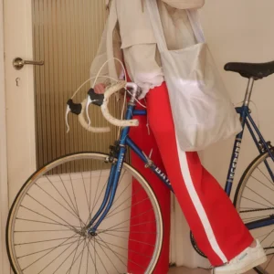 A person in red and white striped trousers, a beige jacket, and white trainers stands indoors next to a blue bicycle with white-taped handlebars, holding the Feel Good Sports Bag - White from a curated Estonian design gift shop. Gate21.ee