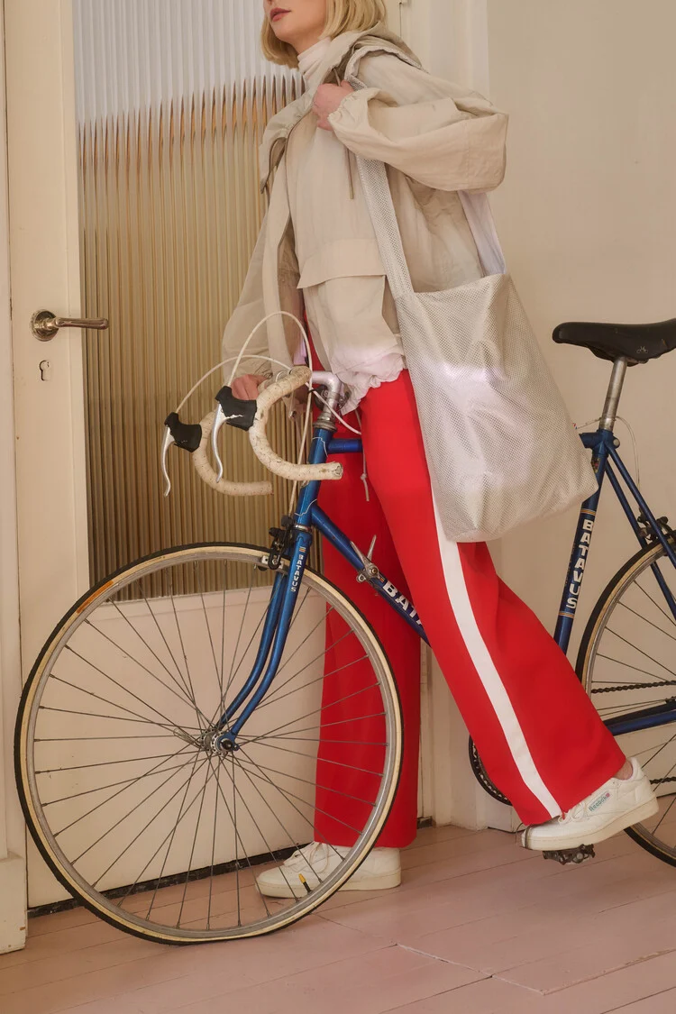A person in red and white striped trousers, a beige jacket, and white trainers stands indoors next to a blue bicycle with white-taped handlebars, holding the Feel Good Sports Bag - White from a curated Estonian design gift shop. Gate21.ee