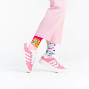A person wears light pink trousers and pink trainers with white stripes, paired with Barbie socks—one showing a cartoon woman’s face, the other featuring the Barbie logo—for a playful look that pops against a clean white background. Gate21.ee
