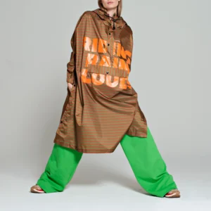 A person stands with legs apart, wearing a brown hooded poncho and wide, bright green trousers from the Breton Green Diary collection—an exemplary display of curated Estonian design against a plain grey background. Gate21.ee