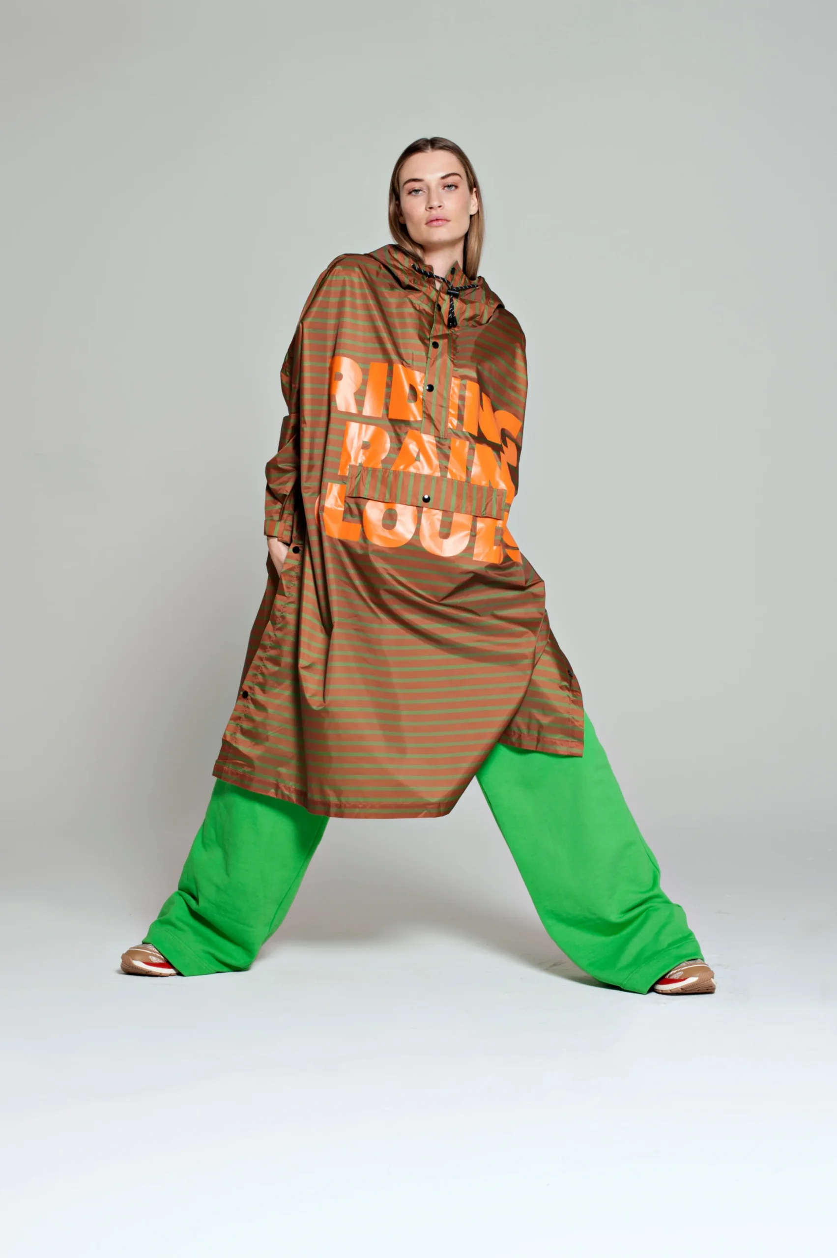 A person stands with legs apart, wearing a brown hooded poncho and wide, bright green trousers from the Breton Green Diary collection—an exemplary display of curated Estonian design against a plain grey background. Gate21.ee