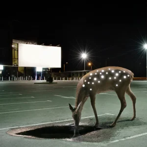 A deer with glowing white spots drinks from a black pool in an empty car park at night, under streetlights and a blank billboard—an unexpected scene that reflects the unique ideas and quiet beauty found in Estonian design. Gate21.ee