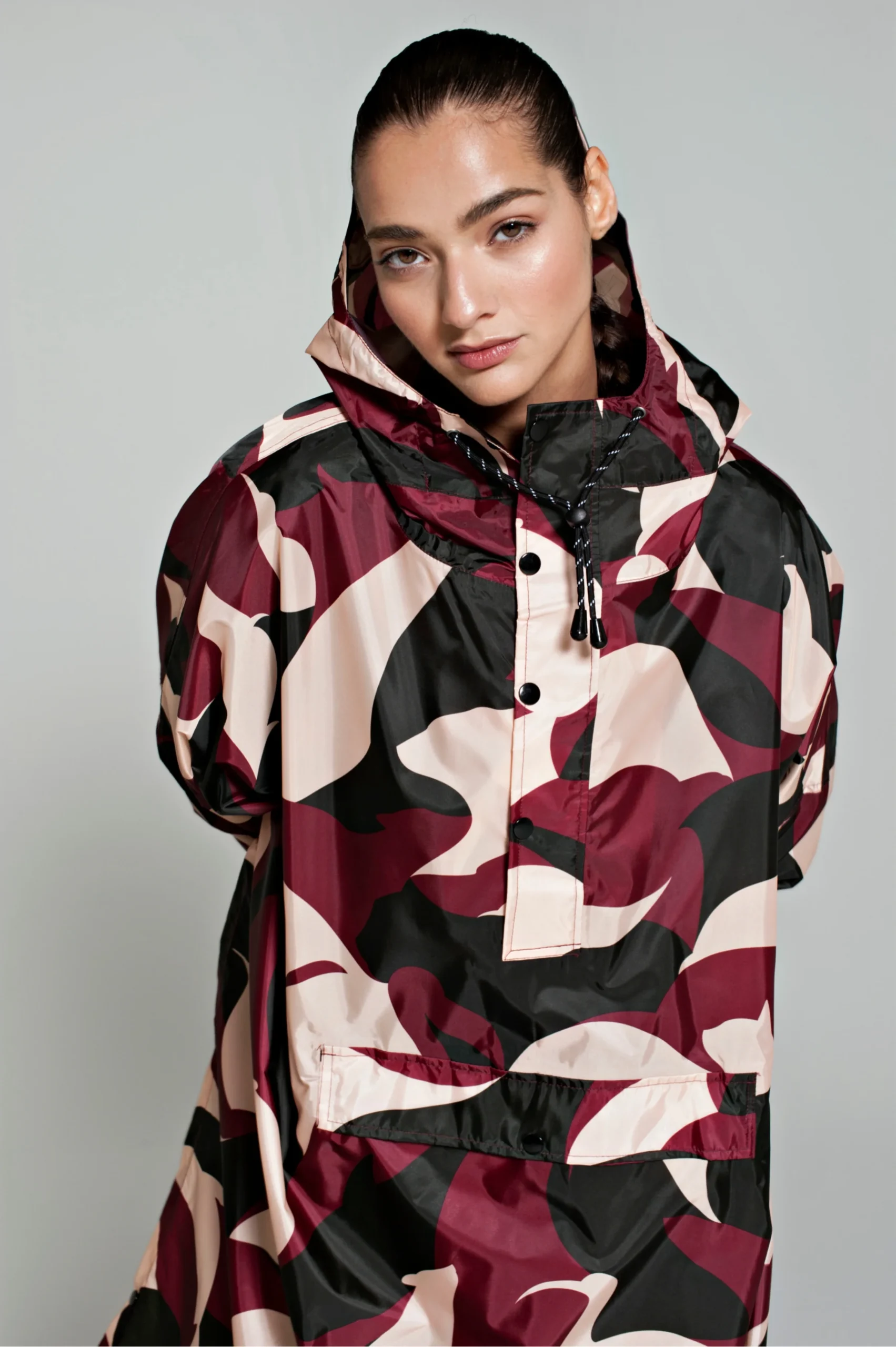 A person with dark hair pulled back wears the Animo Bordeaux hooded jacket, featuring a maroon, black, and beige camouflage pattern—a modern Estonian design—whilst standing against a plain light grey background. Gate21.ee