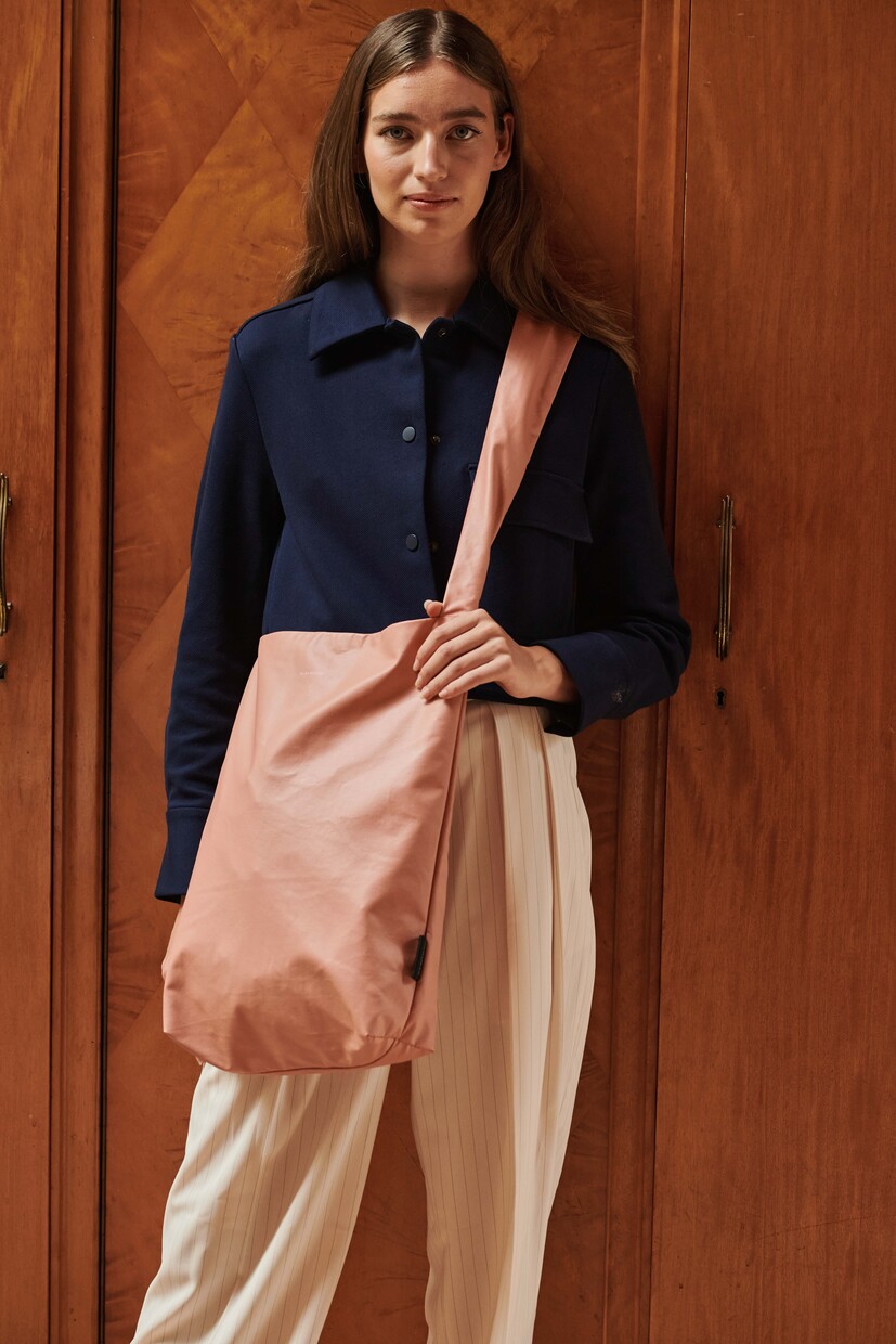 A woman stands indoors by wooden cupboards, wearing a navy blue jacket, white striped trousers, and carrying the Feel Good Bag - Canyon Clay—her style echoing the curated vibe of an Estonian design gift shop. Gate21.ee