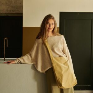 A woman with long hair, dressed in a beige loose jumper and olive green skirt, stands indoors by a counter holding the Feel Good Bag - Prairie Sand—an effortless display of unique and understated Estonian design. Gate21.ee