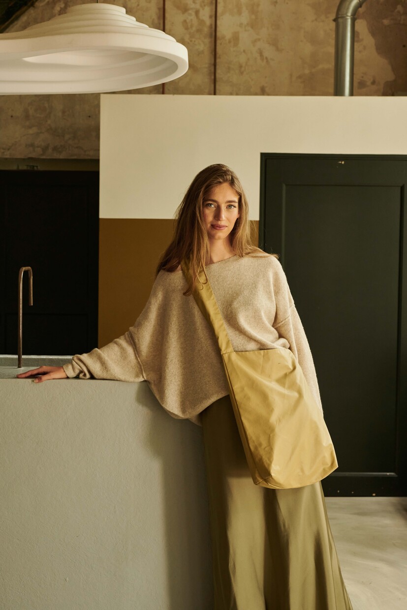 A woman with long hair, dressed in a beige loose jumper and olive green skirt, stands indoors by a counter holding the Feel Good Bag - Prairie Sand—an effortless display of unique and understated Estonian design. Gate21.ee