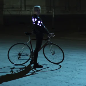 A person with short fair hair stands by a bicycle at night, wearing the “Double - Yellow” jacket—its illuminated circular lights glow in the dimly lit scene, casting long shadows on the pavement and showcasing innovative Estonian design. Gate21.ee