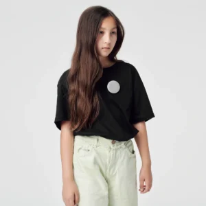 A young person with long brown hair wears the "Single - Silver" black T-shirt, featuring a round, light grey patch on the chest and light-coloured trousers, standing against a plain, light grey background—ideal for Tallinn concept shops. Gate21.ee
