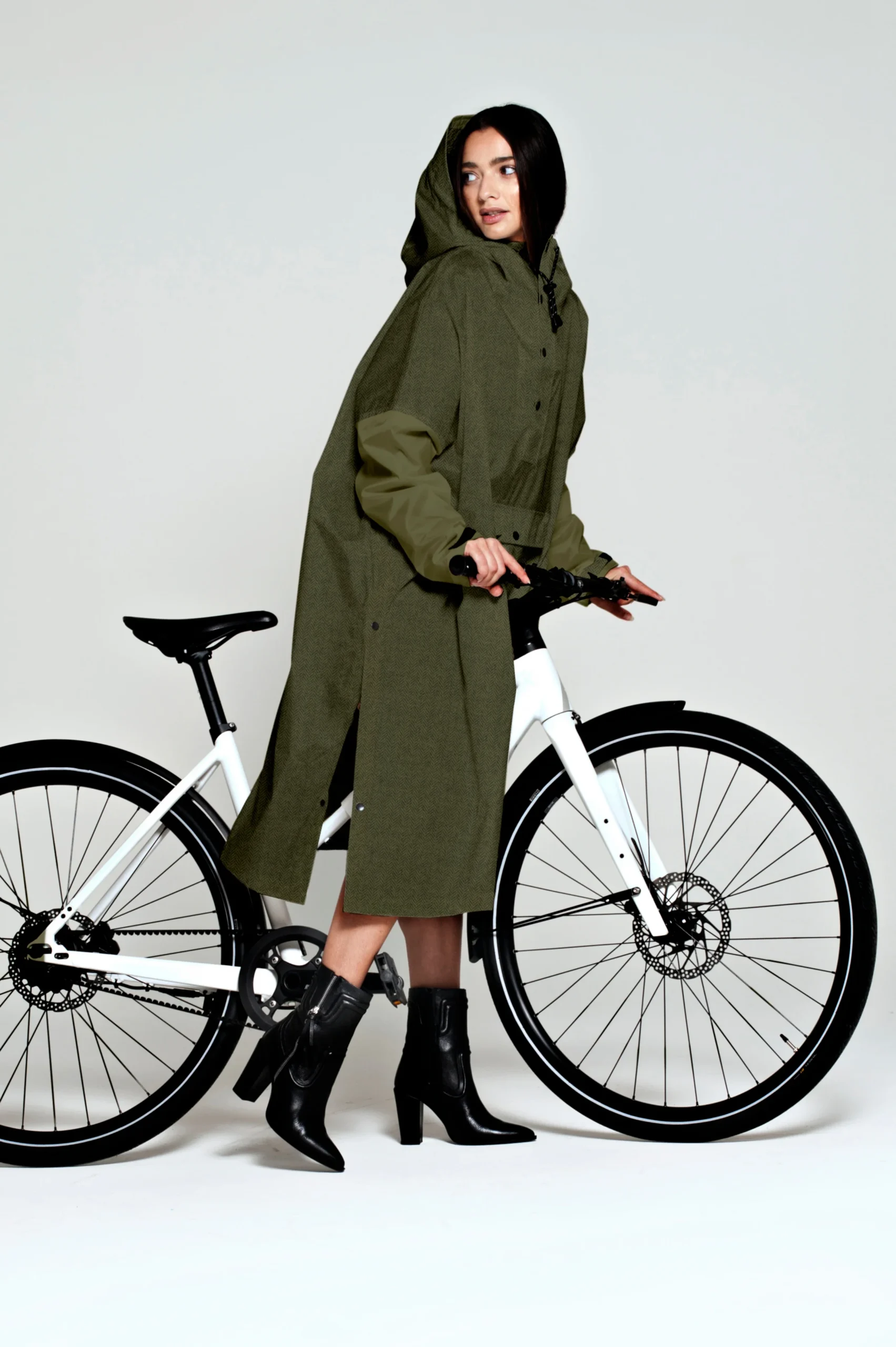 A woman in a long green "Herringbone" hooded coat and black heeled boots stands next to a white bicycle, holding the handlebars, as if she's just left a concept shop full of unique ideas. Gate21.ee