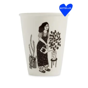 The Plant Lover Cups feature a black illustration of a woman in dungarees watering plants on a white background. This Estonian-designed cup has a blue heart-shaped Bestseller label at the top right. Gate21.ee
