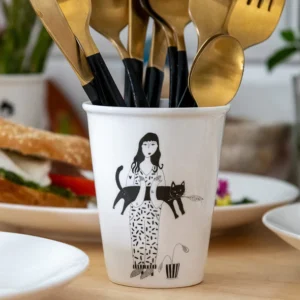 The Black Cat Cups showcase Estonian design with a ceramic cup featuring a woman, black cat, and plant illustration. It holds gold and black cutlery and sits on a wooden table with plates and food in the background. Gate21.ee