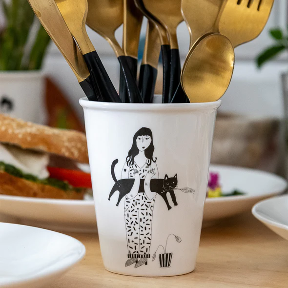 The Black Cat Cups showcase Estonian design with a ceramic cup featuring a woman, black cat, and plant illustration. It holds gold and black cutlery and sits on a wooden table with plates and food in the background. Gate21.ee