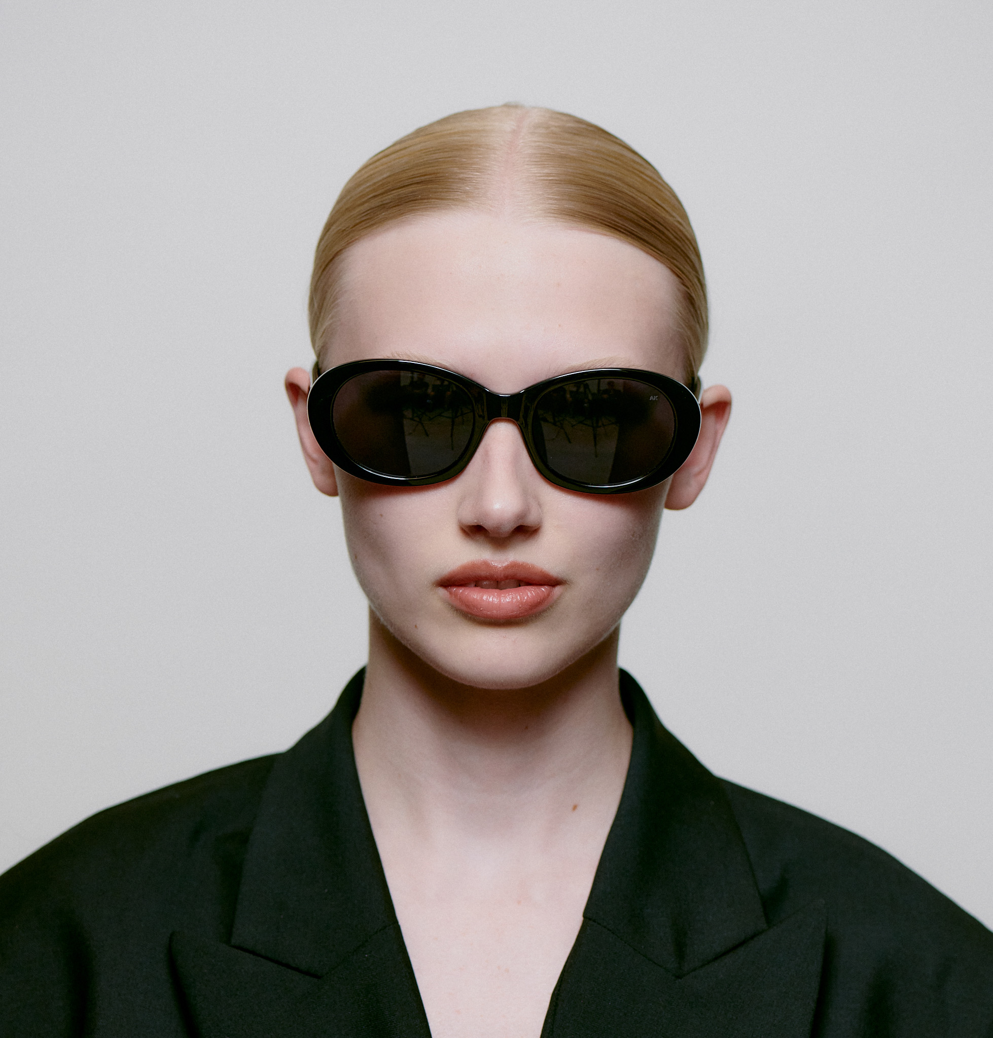 A person with slicked-back blond hair wears the ANMA - Black sunglasses and a dark blazer, gazing directly at the camera against a plain light background—a look inspired by Tallinn’s modern shopping concept stores. Gate21.ee