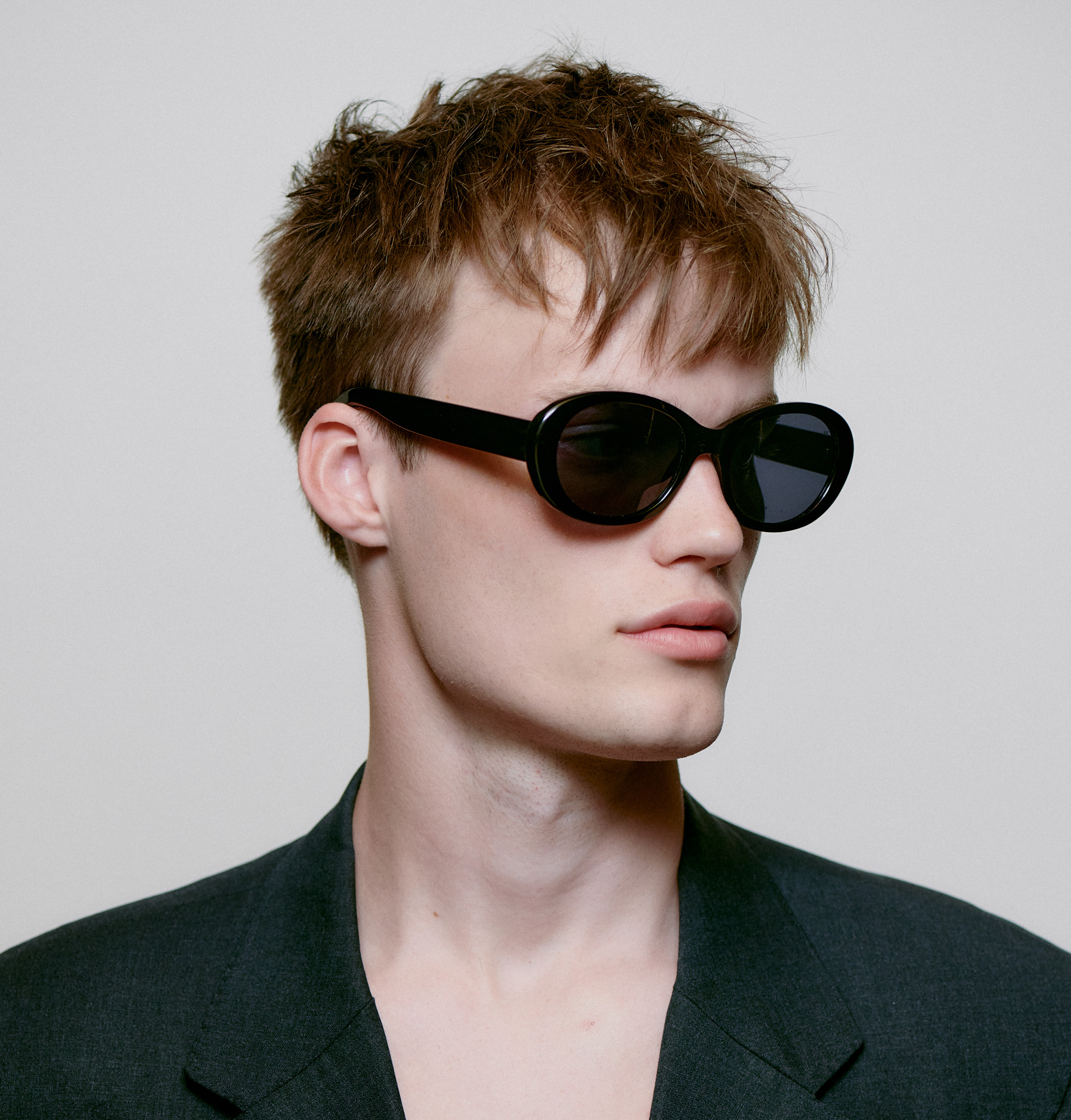A young person with short, tousled light brown hair wears ANMA - Black oval sunglasses and a dark blazer, capturing the chic vibe of a Tallinn concept shop as they glance to the side against a plain light background. Gate21.ee