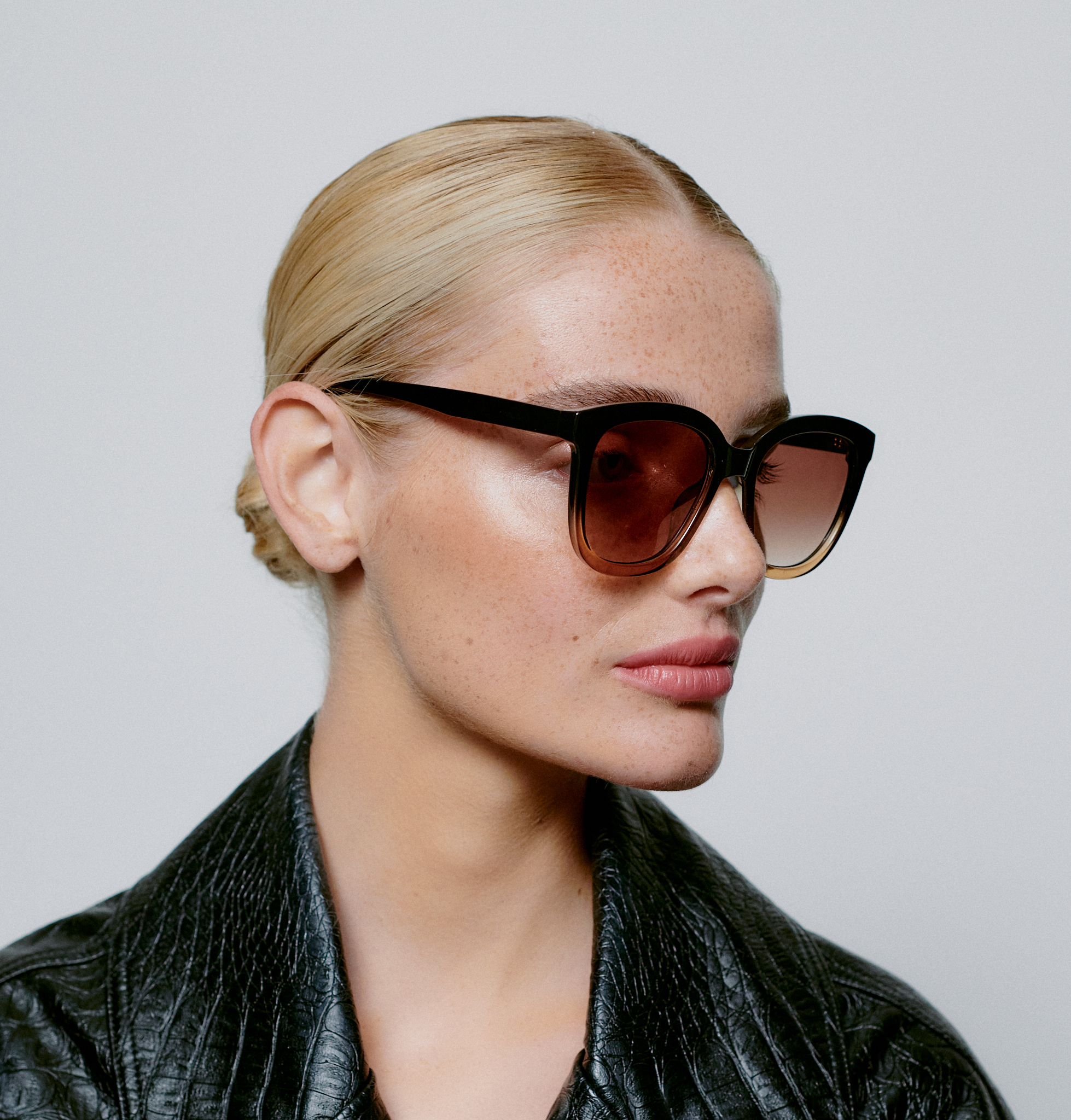 Wearing the BILLY - Black Brown Transparent sunglasses, a light-skinned person with blonde hair pulled back poses in a textured black jacket against a plain background, evoking modern concept store style. Gate21.ee