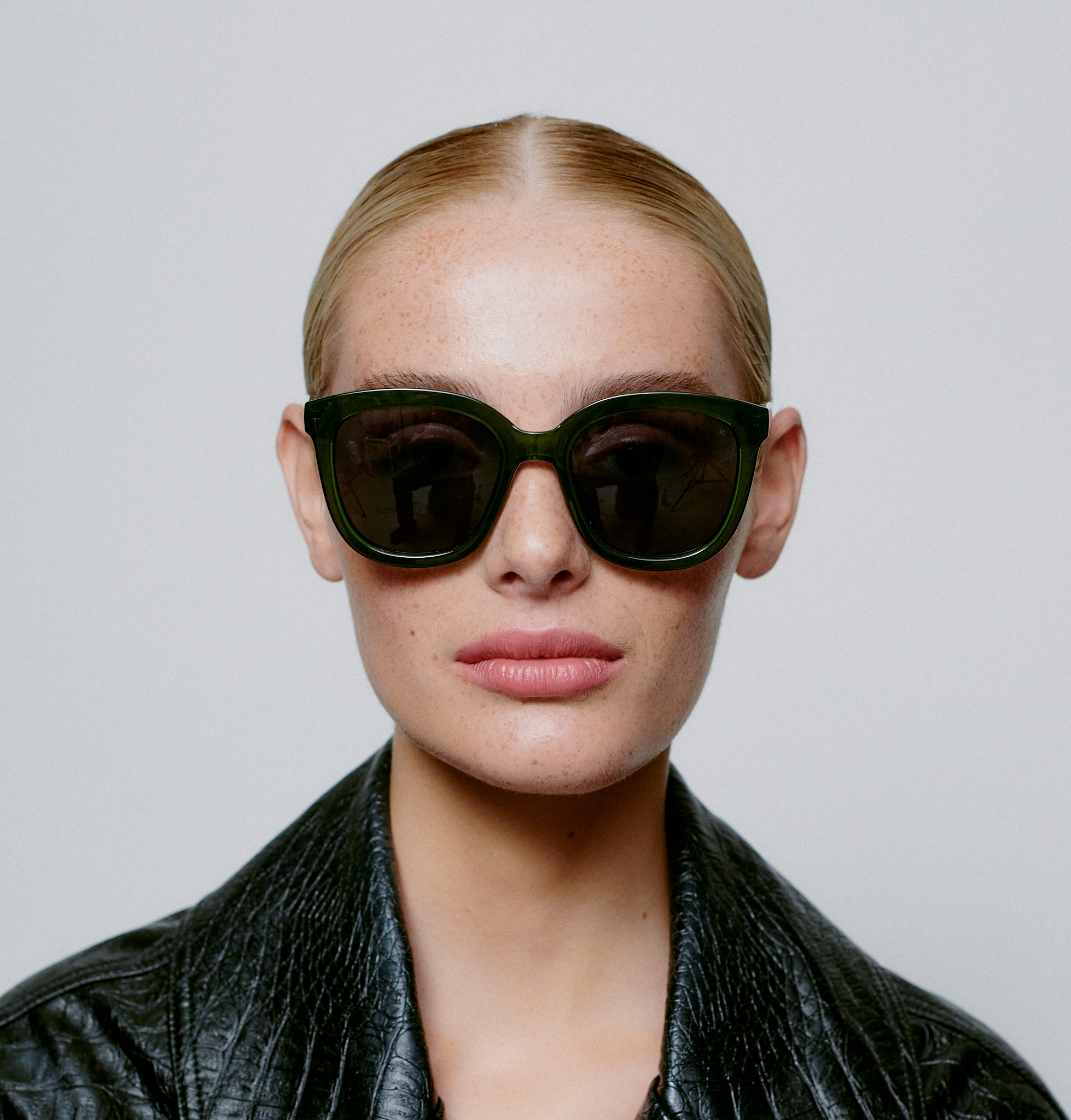 A person with light skin and slicked-back blond hair wears BILLY - Dark Green Transparent sunglasses, embodying the bold spirit of Estonian design as they look directly at the camera against a plain background. Gate21.ee