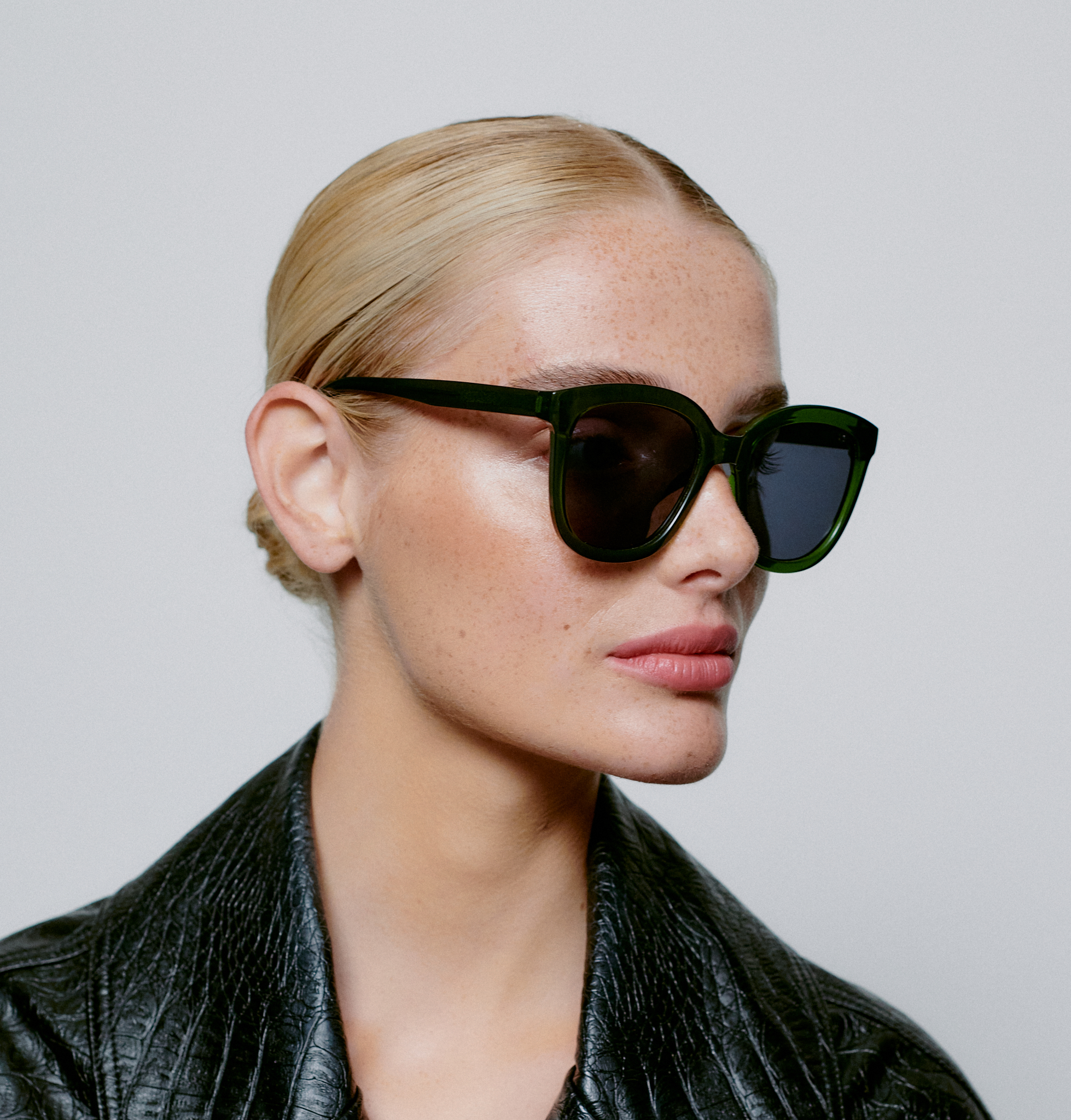 A person with light skin and blonde hair pulled back wears BILLY - Dark Green Transparent sunglasses and a textured black jacket, facing slightly to the left against a plain light background—styled like a curated concept shop. Gate21.ee