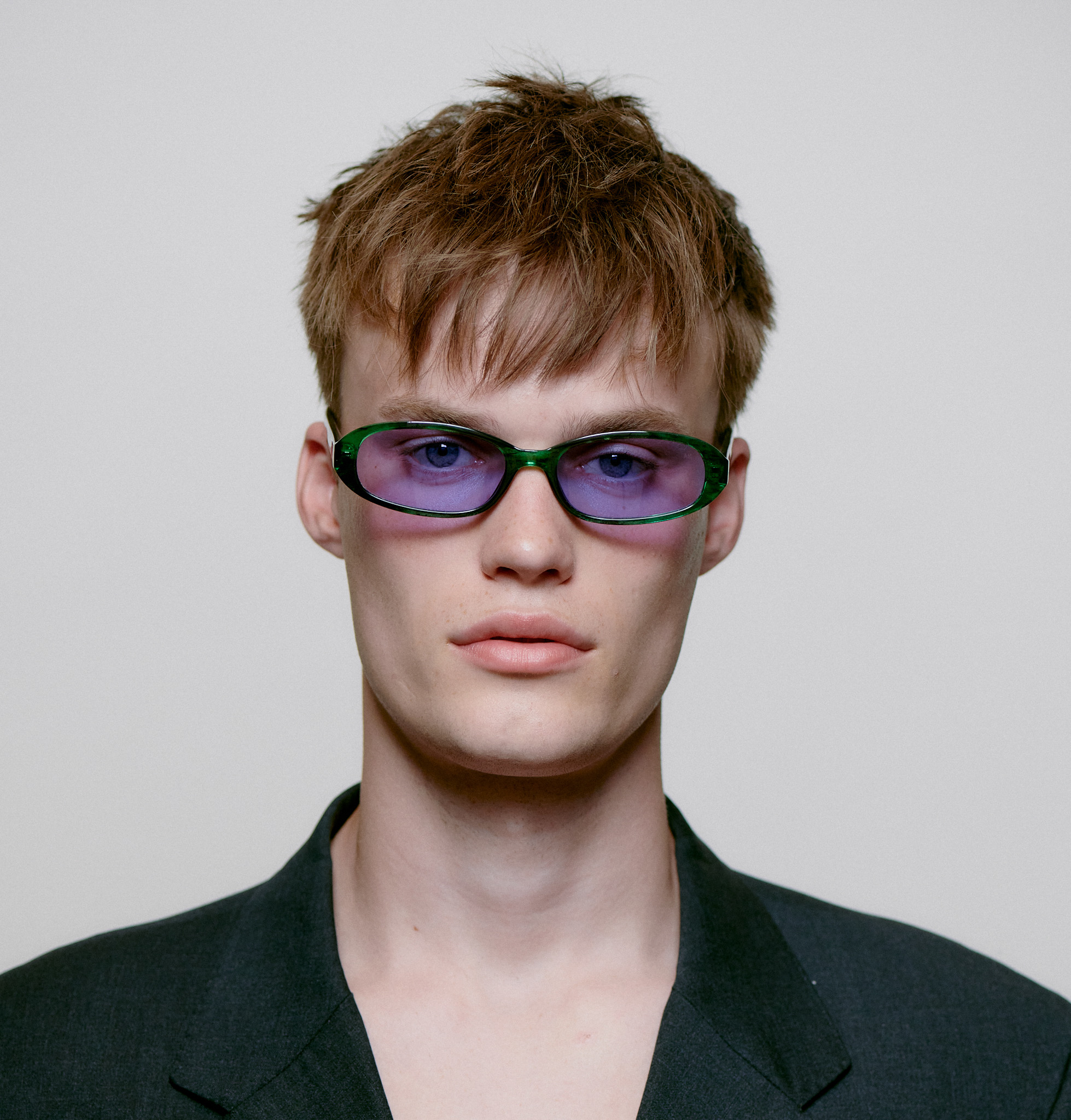 A young person with short, tousled light brown hair wears MACY - Green Marble Transparent sunglasses and a dark blazer, facing forwards against a plain light background—an image inspired by contemporary Estonian design. Gate21.ee