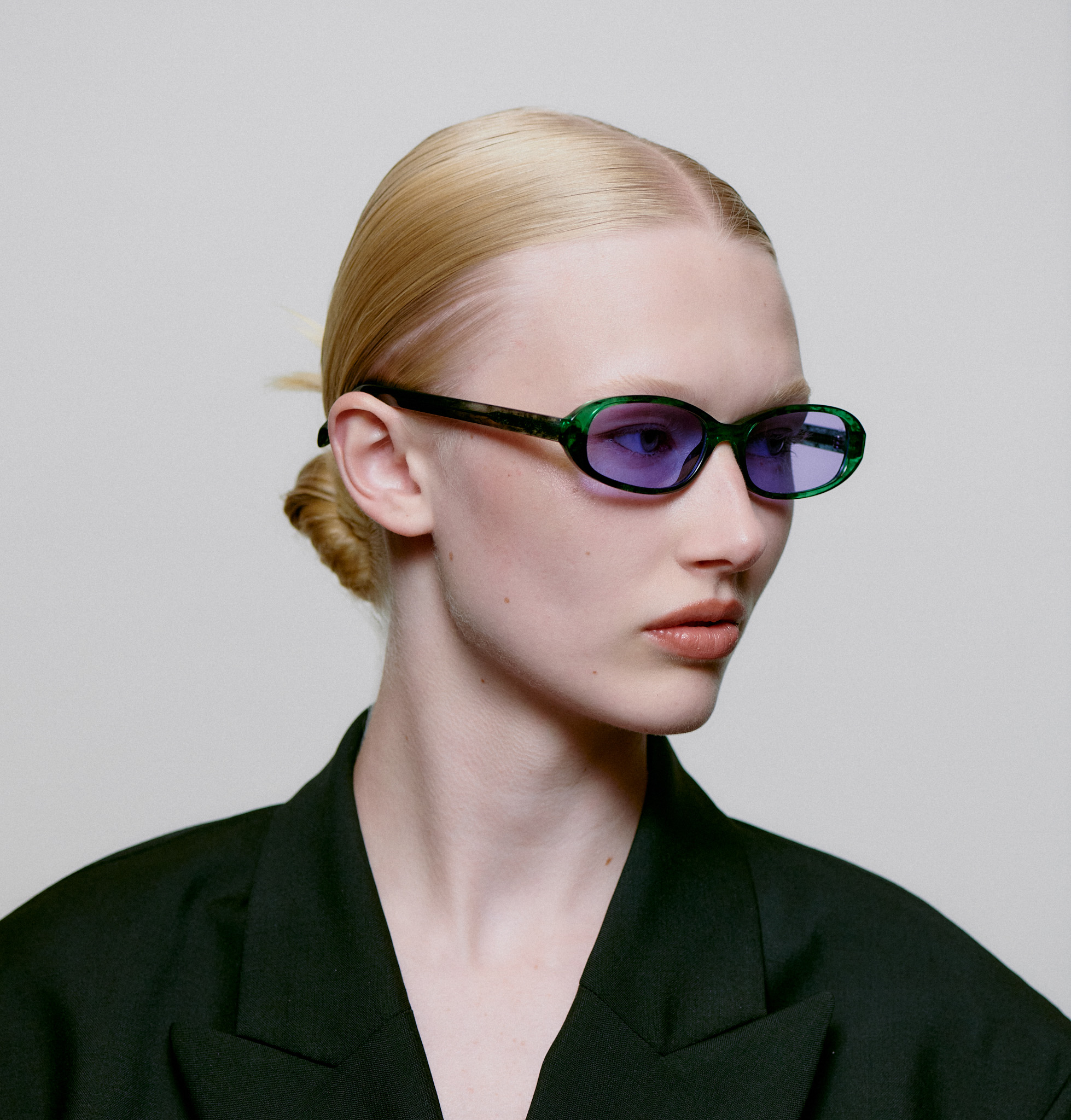 A person with light skin and blonde hair pulled back wears MACY - Green Marble Transparent oval-shaped sunglasses, featuring a green frame and purple lenses, paired with a black blazer against a plain light grey background. Gate21.ee