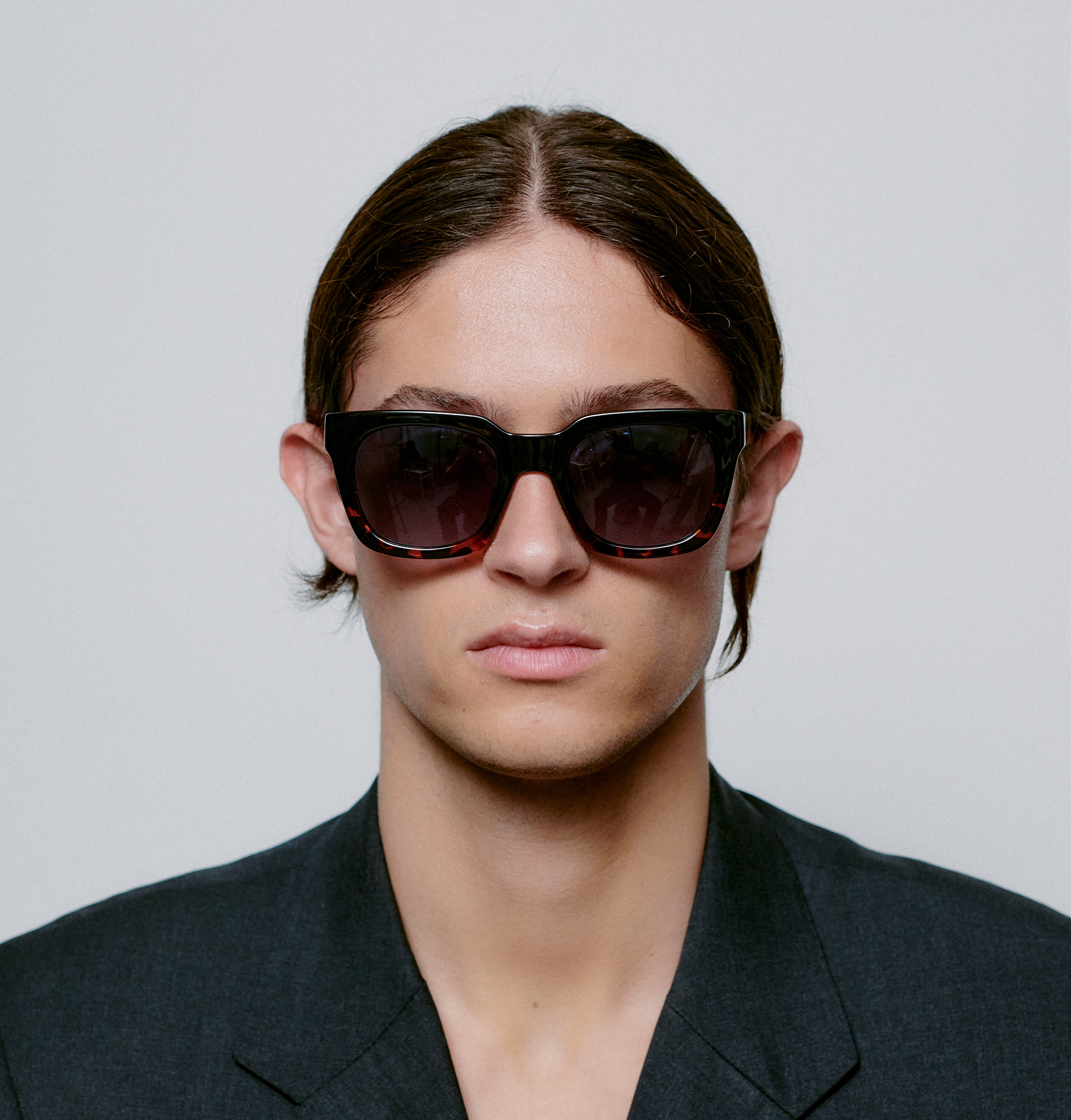 A person with straight brown hair and a centre part wears NANCY - Black Demi Tortoise sunglasses and a dark suit jacket, facing the camera against a plain white background—capturing the curated style of modern concept shops. Gate21.ee