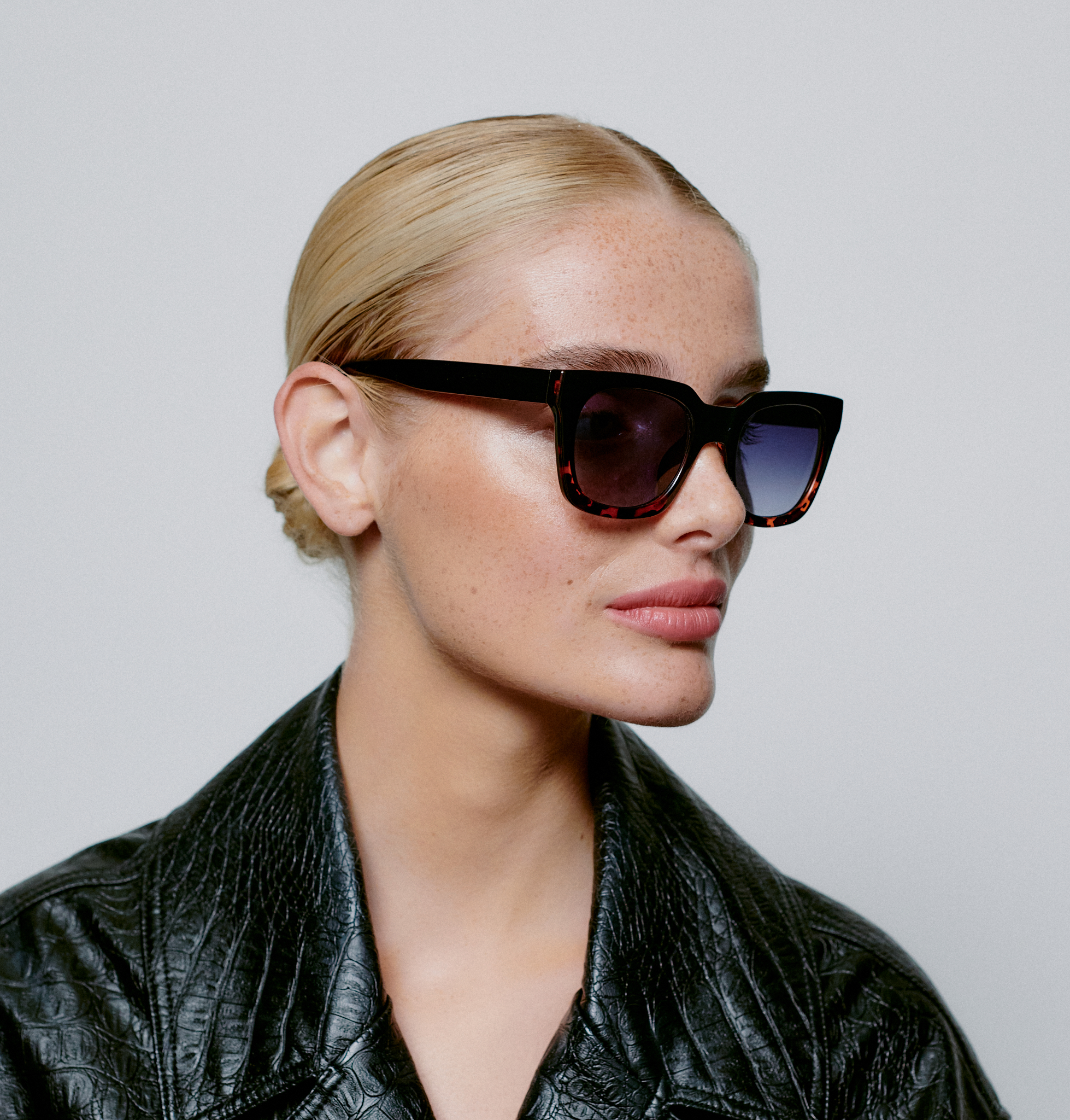 A person with light skin and blonde hair pulled back wears NANCY - Black Demi Tortoise sunglasses and a textured black jacket, facing slightly to the right against a plain grey background. Gate21.ee